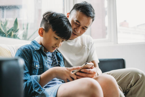 Father and son using a mobile phone together sitting on the sofa at home Father and son using a mobile phone together sitting on the sofa at home. They are spending time together. social media kids stock pictures, royalty-free photos & images