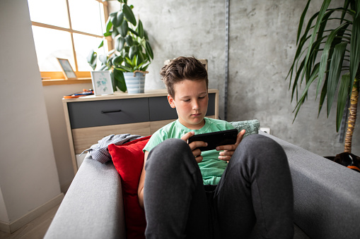 Boy sitting on sofa holding smartphone and playing video games during COVID-19 quarantine at home