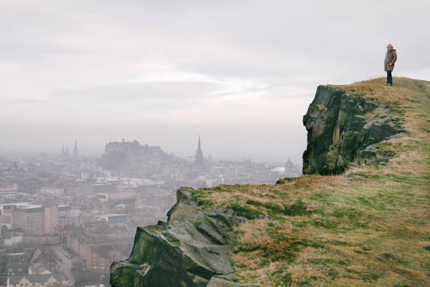 Woman standing on the Arthur's Seat mountain peak in winter in fog with Edinburgh panorama and skyline and Edinburgh Castle visible in the distance stock photo