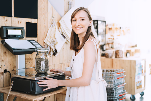 Girl weighs glass jar with hemp seeds. Woman with cotton bag chooses and buys products in zero waste shop. Weighing dry goods in plastic free grocery store. Sustainable shopping at local business