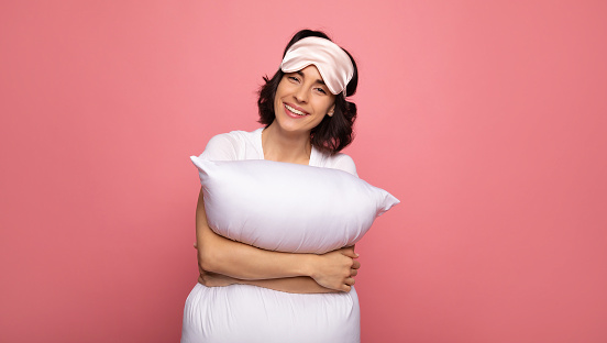 Going to bed. Close-up photo of a happy young woman wearing face mask and hugging a pillow while looking in the camera with a big smile.