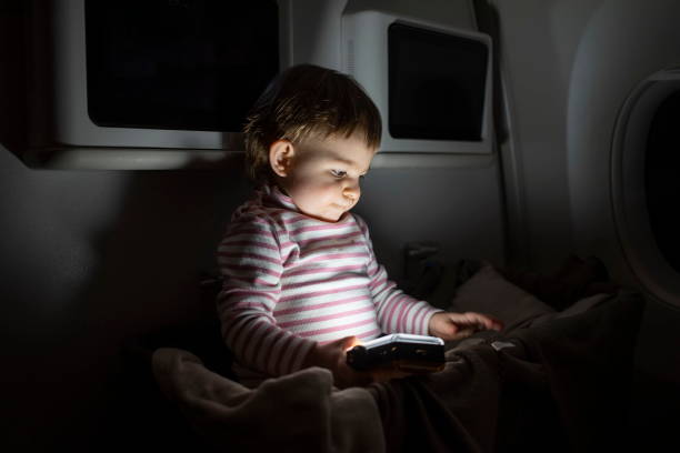 little toddler sitting in special baby bassinet on a plane in the dark and playing with a flashlight stock photo