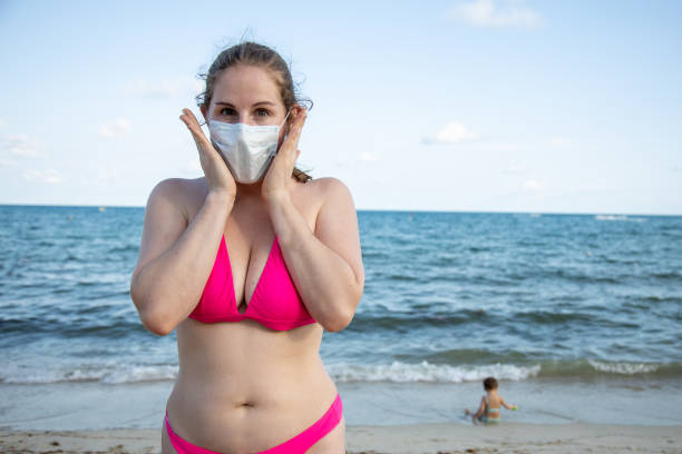 concept of fear of disease while traveling. young caucasian girl in a medical mask and in a swimsuit fearfully looks at the camera. In the background, on the beach by the sea, a child plays in blur. stock photo