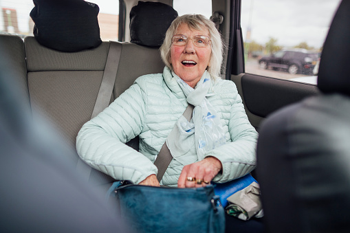 A senior woman sitting in the back of a car zipping her bag up while smiling and looking up. She is getting ready to be driven home.
