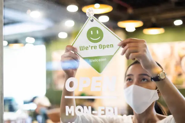 Photo of Business owner Asian woman wear protective face mask ppe hanging open sign at her restaurant / café, open again after lock down due to outbreak of coronavirus covid-19