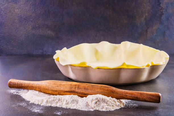 Preparing a pie dough before baking in its pie pan with flour on the worktop and a wooden rolling pin stock photo