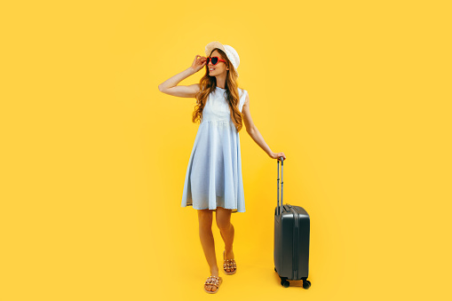 Attractive tourist girl in a summer hat standing with a suitcase isolated on a blue background. Travel, summer vacation concept