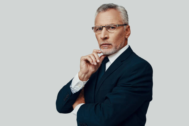 istock Handsome senior man in full suit looking at camera and keeping hand on chin while standing against grey background 1223200217