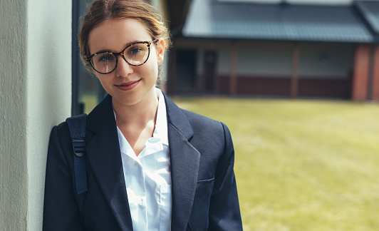 Teenage girl with schoolbag standing in high school campus. Female high school student in uniform looking at camera.