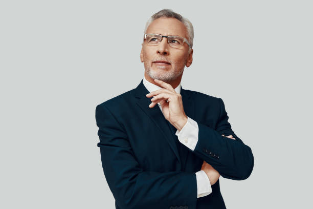 istock Handsome senior man in full suit looking away and keeping hand on chin while standing against grey background 1223201746