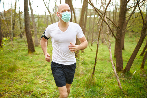 A middle-aged man goes jogging in the forest and stretches while wearing a medical face mask, wearing a white t-shirt and black shorts and a cell phone arm pocket