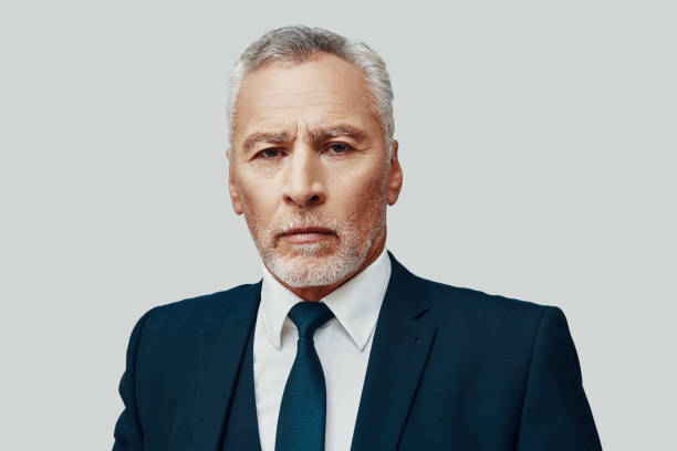 istock Handsome senior man in full suit looking at camera while standing against grey background 1223203947