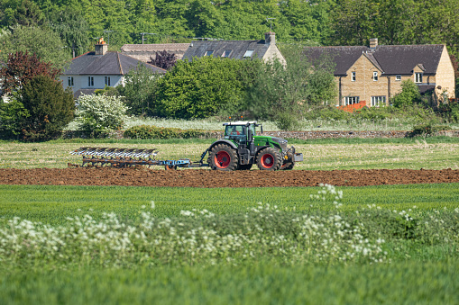 Tractor ploughing a field in The Cotswolds preparing the soil for sowing a crop of Summer wheat. Cottages and houses in the background.