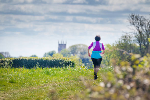 corredora feminina madura solitária fazendo seu exercício diário ao longo de uma pista de campo perto de cirencester, gloucestershire, the cotswolds, reino unido. tiro em um ângulo baixo com um primeiro plano de vegetação rasteira verde bokeh. - bridle path - fotografias e filmes do acervo