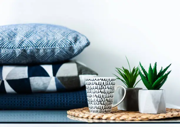 Cozy home interior decor: white and black cup and ornamental plants in pots on a wicker stand on a white table in the room. Blue pillows on background. The quarantine concept of stay home