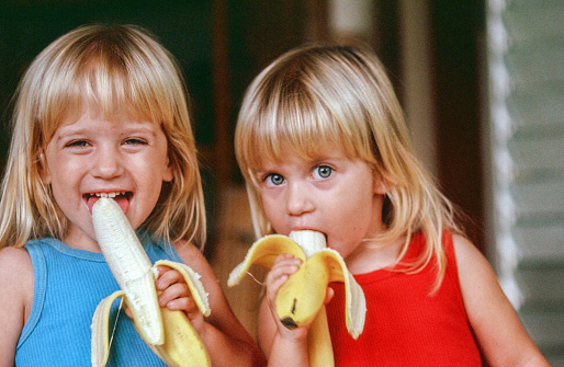 Two young blonde toddlers are smiling at the camera eating bananas. The peel is still partially on the fruit. This horizontal image gives a head and shoulders shot of these girls.