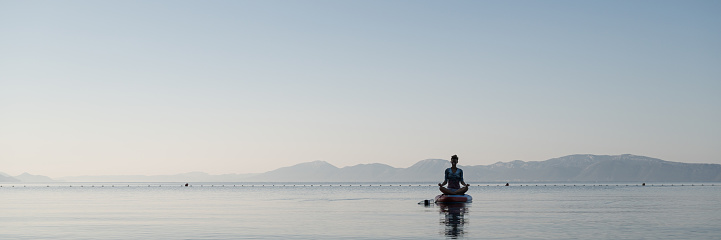 Young woman sitting in lotus position, meditating peaceful on sup board floating on calm morning sea.