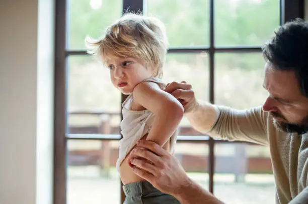 Father with small sick unhappy son indoors at home, checking his hurt back.