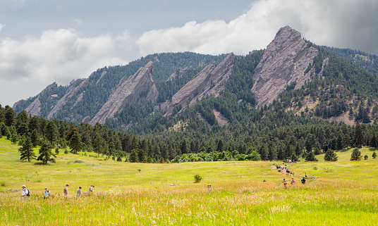 View from Chautauqua Trailhead off Baseline road in Boulder Colorado with hikers on the trail with the Flatirons in the background on a summer day with clouds in the background.