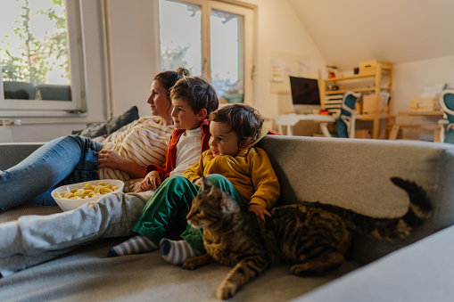Photo of mother and sons watching tv together. Their cat is lying down next to them