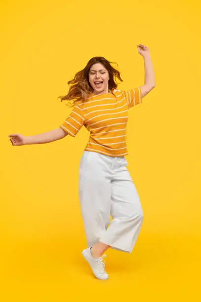 Full body young female in stylish casual outfit energetically dancing with closed eyes against yellow background