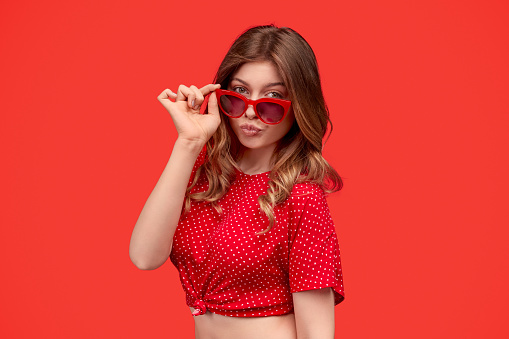Young woman in trendy top pouting lips and looking at camera over shades while standing against red background