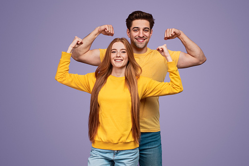 Delighted young man and woman showing biceps and smiling for camera while representing healthy lifestyle against violet background