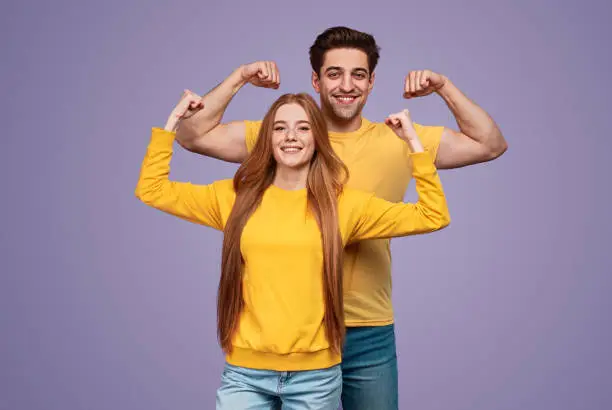 Delighted young man and woman showing biceps and smiling for camera while representing healthy lifestyle against violet background