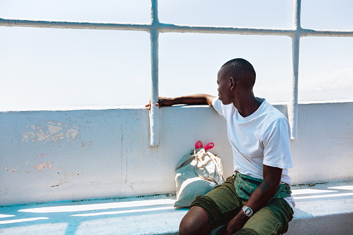 Young african man, 23 years old, wearing shorts and a grey t-shirt, sitting in a tour boat and looking out to the sea, it is near Dar Es Salaam, Tanzania, looking forward to enjoy a day at the beach