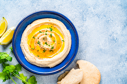 Vegan food: top view of a blue plate with hummus and olive oil shot on bluish tint table. Pita bread, lemon slices and parsley complete the composition. The composition is at the left of an horizontal frame leaving useful copy space for text and/or logo at the right. High resolution 42Mp studio digital capture taken with Sony A7rII and Sony FE 90mm f2.8 macro G OSS lens