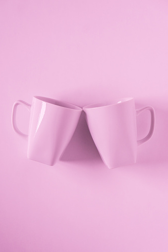 2 monochromatic pink coffee mugs on pink background clinking in cheers with blank empty room space for text, copy, or copyspace. Modern top view concept of two cups with solid background backdrop.