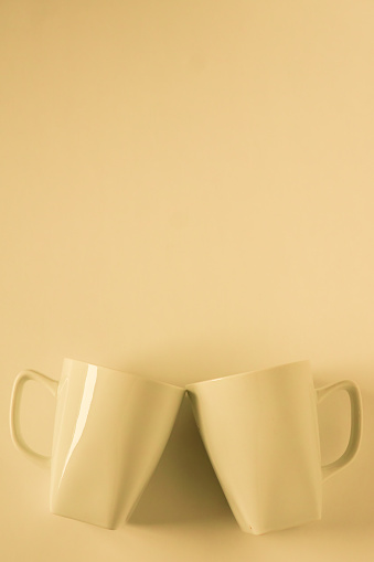 2 monochromatic yellow coffee mugs on yellow background clinking in cheers with blank empty room space for text, copy, or copy space. Modern top view concept of two cups with solid background backdrop.