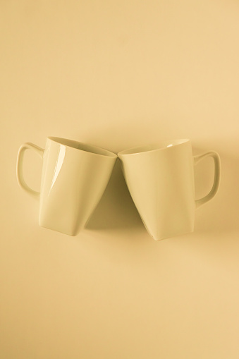 2 monochromatic yellow coffee mugs on yellow background clinking in cheers with blank empty room space for text, copy, or copy space. Modern top view concept of two cups with solid background backdrop.