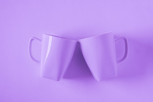 2 monochromatic purple coffee mugs, purplish background clinking in cheers - blank empty room space for text, copy, or copy space. Modern top view concept of two cups with solid background backdrop.