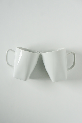 2 monochromatic white coffee mugs on white background clinking in cheers with blank empty room space for text, copy, or copy space. Modern top view concept of two cups with solid background backdrop.