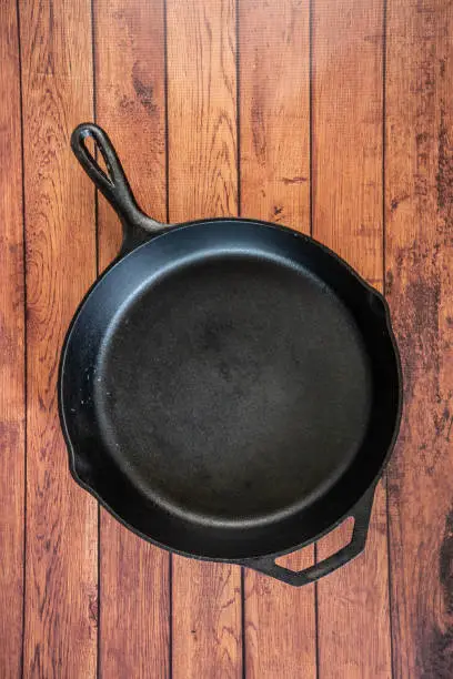 Traditional heavy duty cast iron skillet on wooden surface - isolated top view with copy space. Black cooking utensil - campfire cookware and kitchenware. Kitchen equipment that lasts a lifetime.