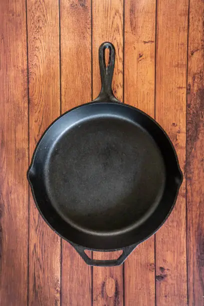 Traditional heavy duty cast iron skillet on wooden surface - isolated top view with copy space. Black cooking utensil - campfire cookware and kitchenware. Kitchen equipment that lasts a lifetime.