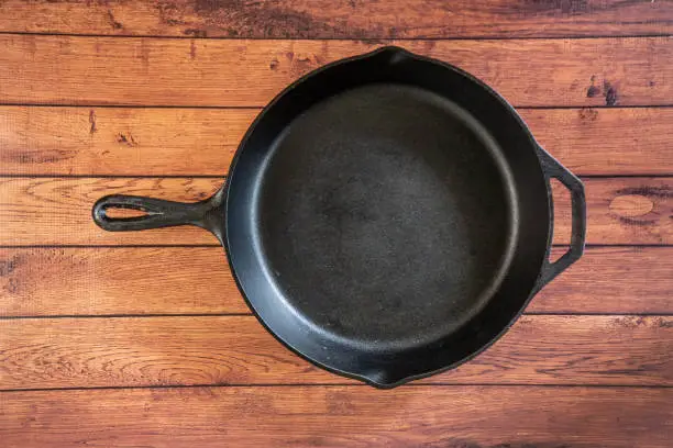 Traditional heavy duty cast iron skillet on wooden surface - isolated top view with copy space. Black cooking utensil - campfire cookware and kitchenware. Kitchen equipment that lasts a lifetime.