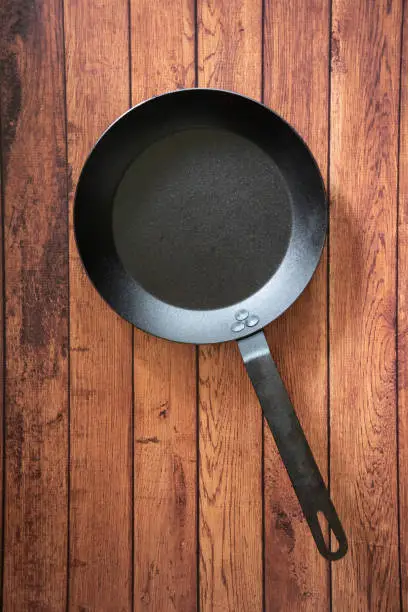 Isolate carbon steel skillet pan on a wooden background - overhead top view of flat lay concept. Professional grade cookware utensil equipment used in restaurants - industry standard for stove top.