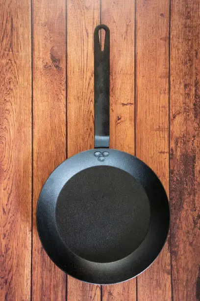 Isolate carbon steel skillet pan on a wooden background - overhead top view of flat lay concept. Professional grade cookware utensil equipment used in restaurants - industry standard for stove top.