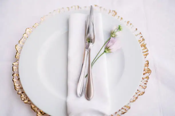 White plate on top of a wildcard with golden edges. In a white plate there is a napkin, a knife, a fork and a purple flower. In the background of the white tablecloth.