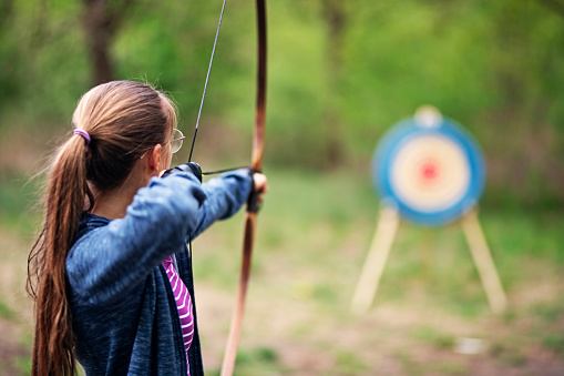 Teenage girl shooting bow outdoors on spring day.\nNikon D850