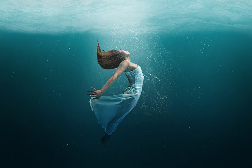 Elegant girl dancer in white dress in a state of levitation under the deep waters of the ocean with sunlight beaming on her face.