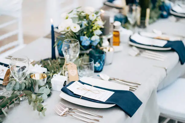 Photo of Wedding dinner table reception. A square plate with a blue cloth towel, knives and forks next to the plate. Flower composition with eucalyptus leaves in the center of the table and burning candles.