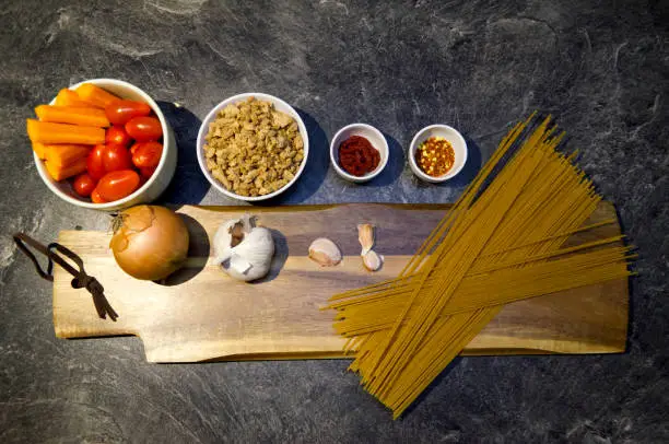 Vegan Wholegrain Spaghetti Bolognese preparation, ingredients, garlic, carrots, tomatoes, soy meat, chili powder on a chopping board