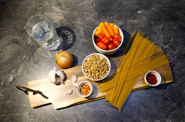 Vegan Wholegrain Spaghetti Bolognese preparation, ingredients, garlic, carrots, tomatoes, soy meat, chili powder on a chopping board