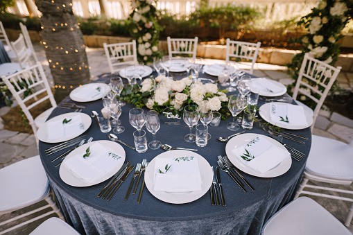 White round plates on a round table with gray tablecloth, white Chiavari chairs with white pillows. A floral arrangement in the center of the table.