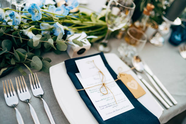 istock Wedding dinner table reception. A square plate with a blue cloth towel, knives and forks next to the plate. Flower composition with eucalyptus leaves in the center of the table and burning candles. 1223293567