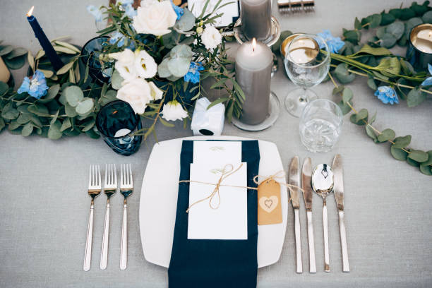 istock Wedding dinner table reception. A square plate with a blue cloth towel, knives and forks next to the plate. Flower composition with eucalyptus leaves in the center of the table and burning candles. 1223295277