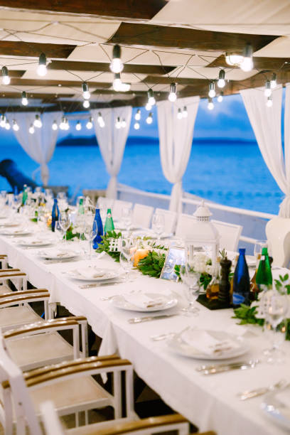 istock Wedding dinner table reception. A long table, against the sea, with a white tablecloth, a garland with incandescent light bulbs over it. Wooden white chairs. Flower composition in the center of table 1223296633